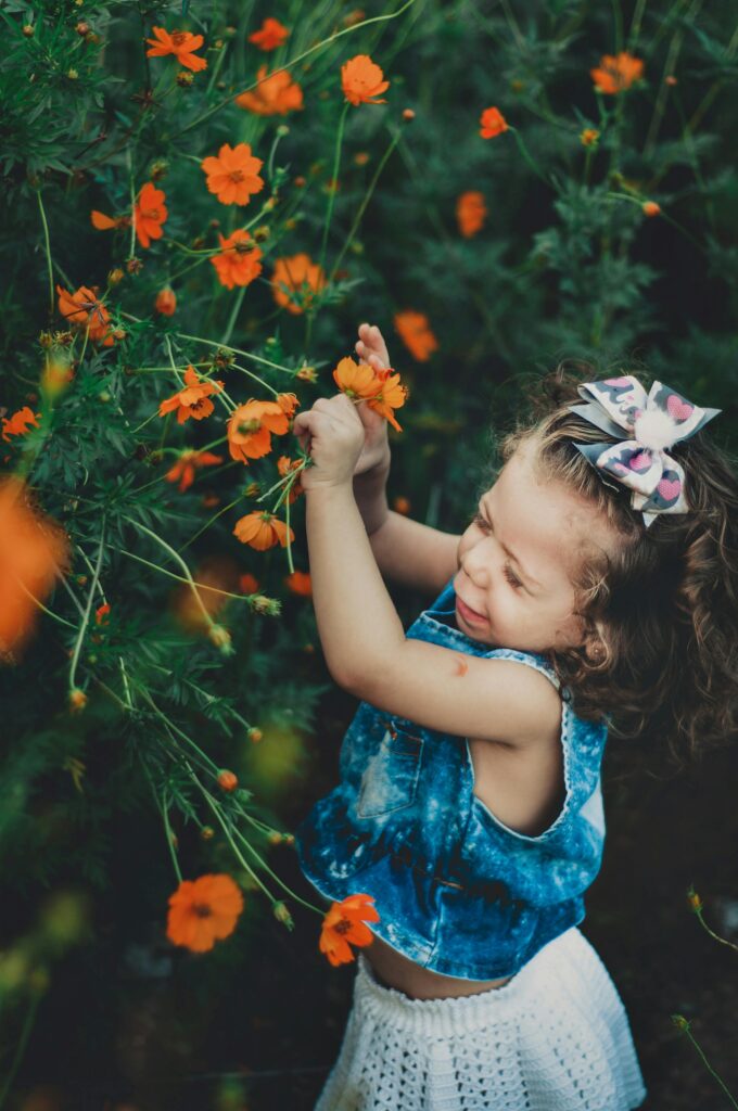 Side view of adorable capricious little girl in casual outfit with bow picking red blossoming flowers